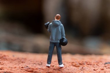 Minyatür insanlar, dambılları kaldıran bir adam desenli bir arka plana ayarlıdır. Gri kapüşonlu ve koyu renk pantolonlu figür, minyatür bir dünyada forma ve güce odaklanmayı temsil ediyor..