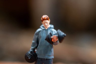 Minyatür insanlar, dambılları kaldıran bir adam desenli bir arka plana ayarlıdır. Gri kapüşonlu ve koyu renk pantolonlu figür, minyatür bir dünyada forma ve güce odaklanmayı temsil ediyor..