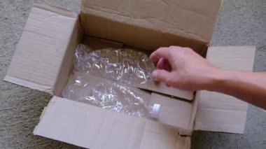 İnsan elinin buruşmuş plastik şişeleri karton bir kutuya koyarkenki yakın çekimi. Açı, kutunun içinden bir görüntü veriyor. Elin hareketine odaklanıyor. Dikkatlice şişeleri düşürüyor..