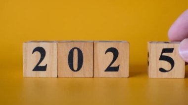 Ahşap blokları siyah sayılarla ayarlayan bir elin yakın çekimi 2025 'i canlı sarı bir arka plana karşı oluşturur.