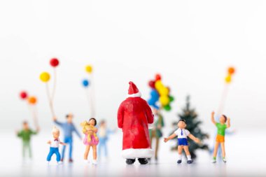 Minyatür Noel Baba elinde çocuklarla ve renkli balonlarla çevrili bir hediyeyle arka planda küçük bir Noel ağacı ile.