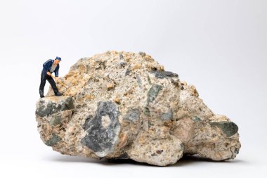 Beyaz arkaplandaki büyük bir kayayı parçalamak için kazma kullanan minyatür bir işçi.