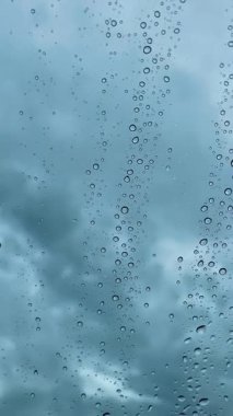 Yağmurlu bir günde, cam bir pencerenin üzerinde serin ve karamsar bir yağmur damlası görüntüsü. Arka plandaki bulutlu gökyüzünün yumuşak mavi tonları