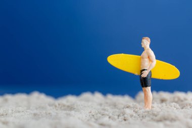 Sarı sörf tahtası tutan bir sörfçünün minyatür bir figürü. Saf mavi arka planı olan ince bir kumun üzerinde duruyor.