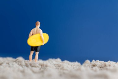 Sarı sörf tahtası tutan bir sörfçünün minyatür bir figürü. Saf mavi arka planı olan ince bir kumun üzerinde duruyor.
