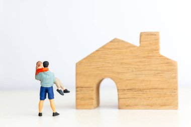 Minyatür bir adamın, tahta bir ev figürünün yanında bir kadını kollarında taşıdığı garip kavramsal tasviri.