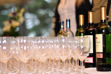 Many wine glasses in rows and different wine bottles on a table with a white tablecloth. Restaurant setting or an event like drink or wine tasting. Bokeh background and people in the back.