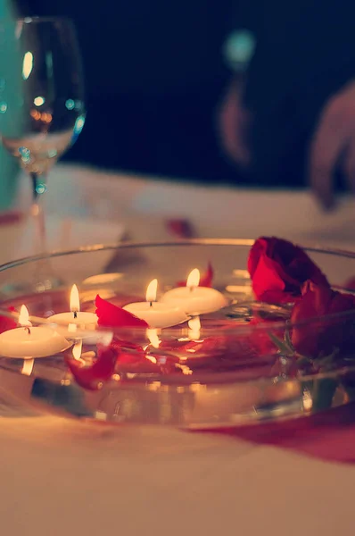 Candles or tealight candles floating in water with red roses and rose petals in a shallow glass dish or bowl. Decoration on a romantic restaurant table, wine glass in the background.