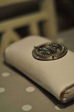 White leather purse or big wallet with brown leather borders, zipper or zip and a metal detail piece. Purse or womens wallet on a table with a chair in the background.