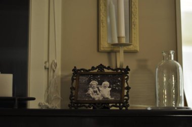 Old picture of children, kids or siblings in intricate wooden or metallic picture frame on top of a wooden dresser, chest or drawer. Around it a glass vase or bottle, candles and a mirror on the wall.
