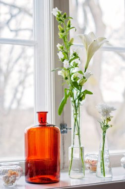 Many glass vases and jugs and flowers on a windowsill. Clear glass vases and one orange glass vase with different white flowers like lilies in front of a window.