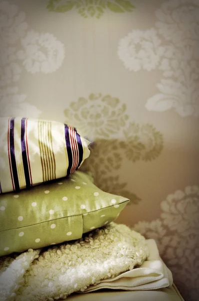 Pile of pillows and blankets on the bed or on the couch or sofa. Stacked pillows in many colors and patterns like white, green, polka dot and striped. Neutral colored wallpaper in the background.
