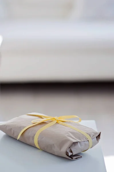 Present wrapped in brown wrapping paper and tied with a yellow ribbon. Gift waiting on a white table with a white couch or sofa in the background in the living room.