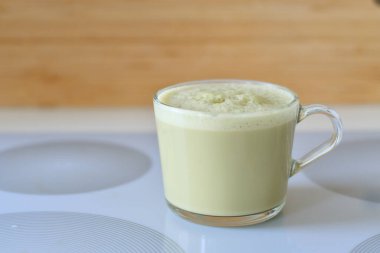 Matcha latte with oatmilk in a glass mug or cup with a neutral and blurry background. Matcha latte is yellow or green in color and has foamy and frothy oatmilk.
