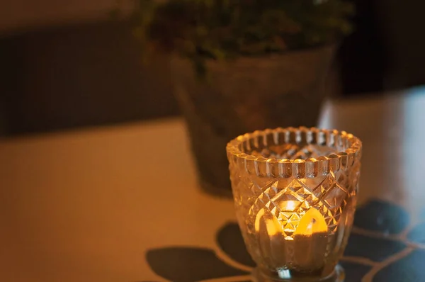 Lit tealight candle in an intricate mosaic-like glass cup or candle holder on the table. Green potted houseplant in the background. Small candle flame burning brightly.