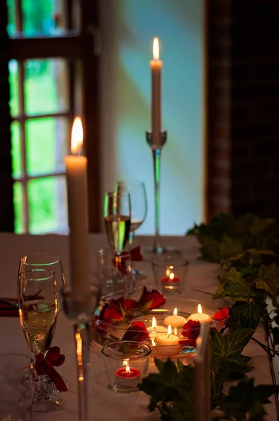 Decorated Christmas table with candles, candlesticks, tealight candles, candle holders, artificial plants, white tablecloth and wine glasses. Romantic dim lighting, holiday vibes.