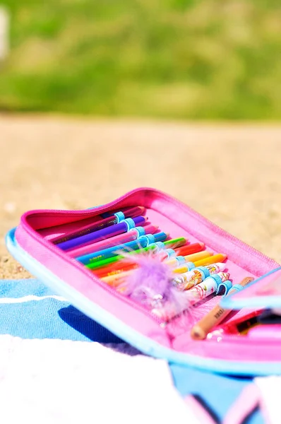 Pink and blue pencil case filled with colored pencils and other craft supplies on a blue beach towel on the beach. Green forest area in the background and warm sand on a sunny summer day.