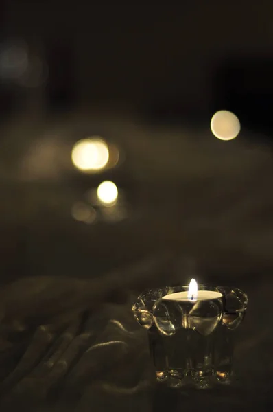 Close-up of an arrangement of candles on a table with white fabric cloth. Tealight candles in clear glass cups in dim lighting and romantic, ambient feeling. Small flames burning, blurry background.