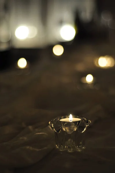 Close-up of an arrangement of candles on a table with white fabric cloth. Tealight candles in clear glass cups in dim lighting and romantic, ambient feeling. Small flames burning, blurry background.