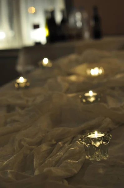 Arrangement of candles on a table surrounded by white fabric. Tealight candles in clear glass cups in dim lighting and romantic, ambient feeling. Small flames burn brightly.