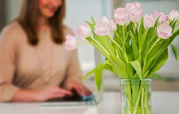 Working alone at home office, pink Spring tulips on a white clean table and kitchen. Happy working woman at home on a laptop computer