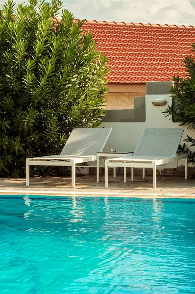 Two white beach chairs next to a pool with warm blue water. Hotel pool setting. Big green bush plant and roof of a house in the background. Lovely summer vacation in Greece.