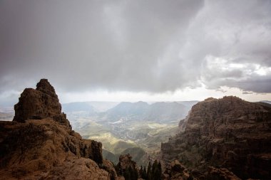 İspanya, Gran Canaria 'daki dağların ve volkanik kayalık tepelerin çarpıcı manzarası. Gökyüzü bulutlu ve puslu. Dağlar arasındaki vadide birçok ev, bina ve yol olan bir kasaba var..
