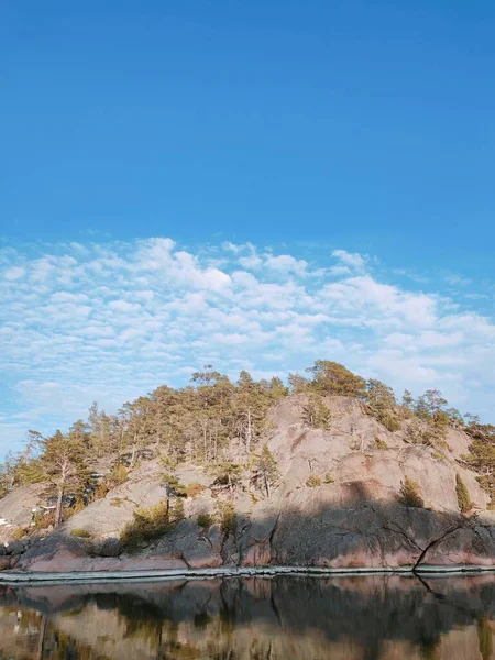 大海中的一个大石头岛或芬兰的一个湖泊 它是完全岩石 有针叶树 如松树和一些灌木 小岛反射在水面上 蓝蓝的天空闪烁着柔和的云彩 — 图库照片