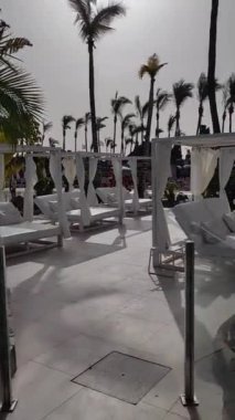 Havuz çevresinde yürüyüş ve güzel turkuaz su. Uzun palmiye ağaçları ve diğer yeşil bitkiler güçlü rüzgarda sallanıyor. Havuzun etrafında sandalyeler ve güneş yatakları. Sıcak ve güneşli ama rüzgarlı bir gün..