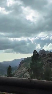 İspanya, Gran Canaria 'daki dağlarda araba sürmek. Arabanın camından bakınca dağlar, evler ve ağaçlar görünüyor. Manzara yanıp sönüyor. Gökyüzü bulutlu, ama güneş bulutların arasından görünüyor..
