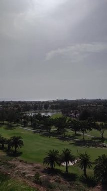 Golf sahasında golf sahası, tepeden atılan sığınaklar ve ağaçlar. Golf sahasındaki golfçüler. Arkasında birçok bina ve ev olan bir şehir manzarası var. Mavi gökyüzü ve ufuktaki deniz kıyısı.