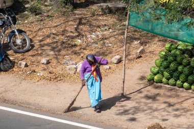 Hassan, Karnataka, India - January 9 2023: An Indian woman sanitation worker sweeping a street with a broom. clipart