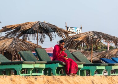 Calangute, Goa, Hindistan - Ocak 2023: Calangute sahilindeki bir kulübede tek başına oturan kırmızı sari giyen Hintli bir kadının samimi portresi.