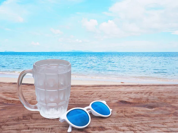 在蓝色天空和白云背景的夏季海滩上方的木制桌面上 关上一杯冷水 — 图库照片