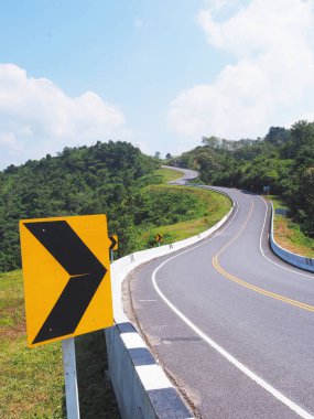 Dağdaki tehlikeli eğri yolda siyah ve sarı yönlü uyarı oku işareti.