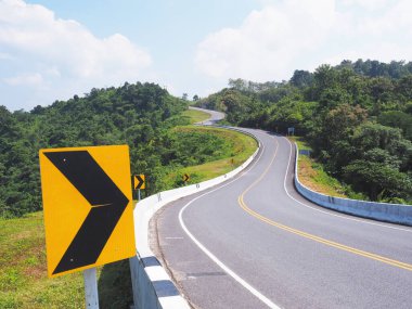 Dağdaki tehlikeli eğri yolda siyah ve sarı yönlü uyarı oku işareti.