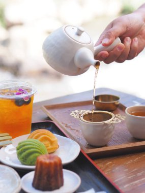 El ele tutuşmuş çaydanlık sıcak çayı bir fincana döküyor ve kek Japon kafesindeki ahşap bir masada. Öğleden sonra çay molası..