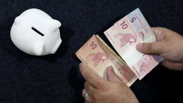 Masasında Domuz Kumbarası Olan Brezilya Banknotlarını Sayan Bir Adamın Elleri — Stok video