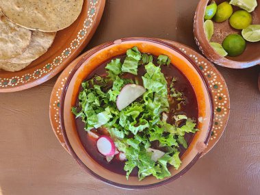 Baharatlı kırmızı pozole tabağı, yanında marul, tost ve limon.