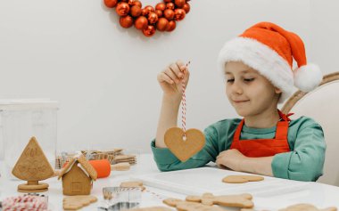 Noel Baba şapkalı şirin bir çocuk mutfakta otururken zencefilli kurabiyeleri ipte tutuyor. Yeni yıl arifesi ve Noel arifesi.