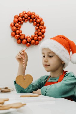 Noel Baba şapkalı şirin bir çocuk mutfakta otururken zencefilli kurabiyeleri ipte tutuyor. Yeni yıl arifesi ve Noel arifesi.