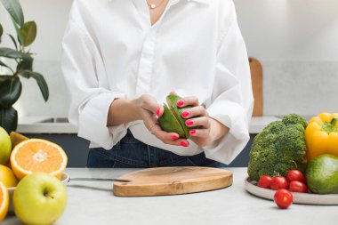 Genç bir kadın taze meyve ve sebzelerle çevrili mutfak masasında olgun bir avokadoyu ikiye böler. Vejetaryenlik kavramı, sağlıklı beslenme.