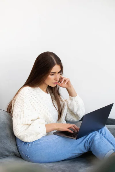 stock image An attractive brunette woman is sitting on the couch at home and studying information on the Internet. The woman looks at the laptop lying on her lap. Online training. Remote work.