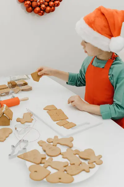 サンタの帽子の男の子は台所のテーブルの上に生姜クッキーを置く 大晦日とクリスマス — ストック写真