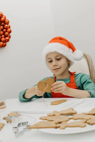 サンタの帽子のかわいい男の子は クリスマスの装飾で飾られたキッチンで心臓の形をしたジンジャークッキーの穴にロープを伸ばします クリスマス休暇の概念 — ストック写真