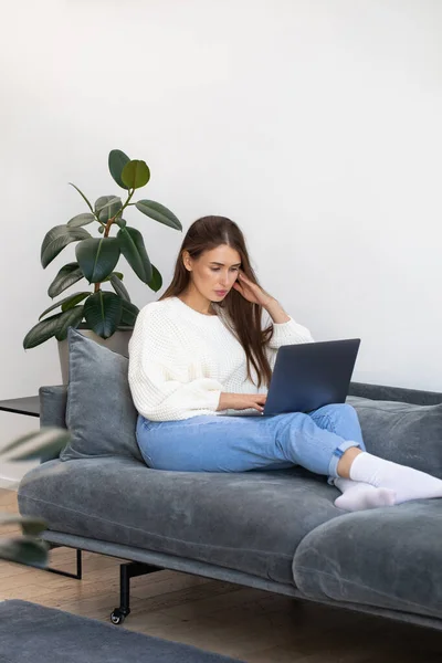 An attractive freelance woman is sitting on the couch and using a laptop to work online. The concept of remote work, training, online shopping.