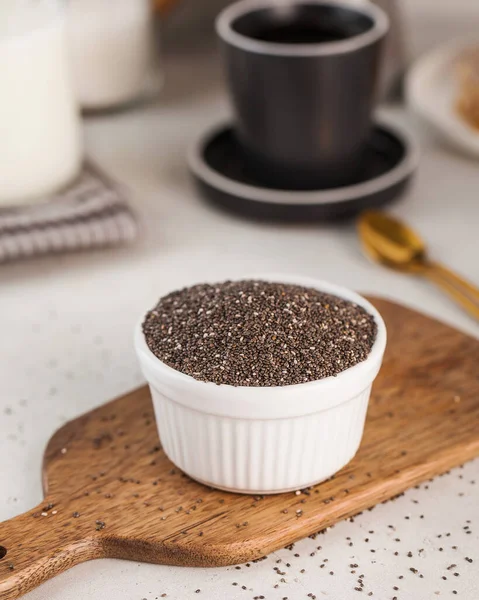 stock image Chia seeds in a white bowl on a wooden board in the background a coffee cup. Superfood concept.