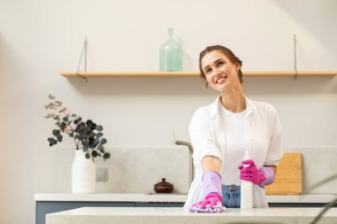 Lastik eldivenli çekici genç bir kadın bir şişe deterjan tutuyor ve gülümseyerek mutfaktaki tezgahı siliyor. Evi temizleme fikri. Temizlik ürünleri.