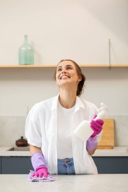 Lastik eldivenli çekici genç bir kadın bir şişe deterjan tutuyor ve gülümseyerek mutfaktaki tezgahı siliyor. Evi temizleme fikri. Temizlik ürünleri.