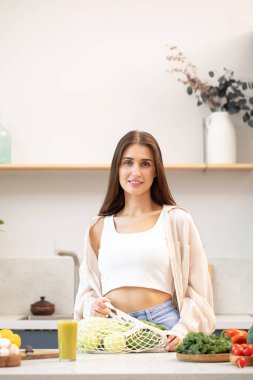 Bir çanta dolusu taze sebze ve meyveli çekici genç bir kadın mutfakta duruyor. Sağlıklı beslenme, yaşam tarzı ve vejetaryenlik kavramı.